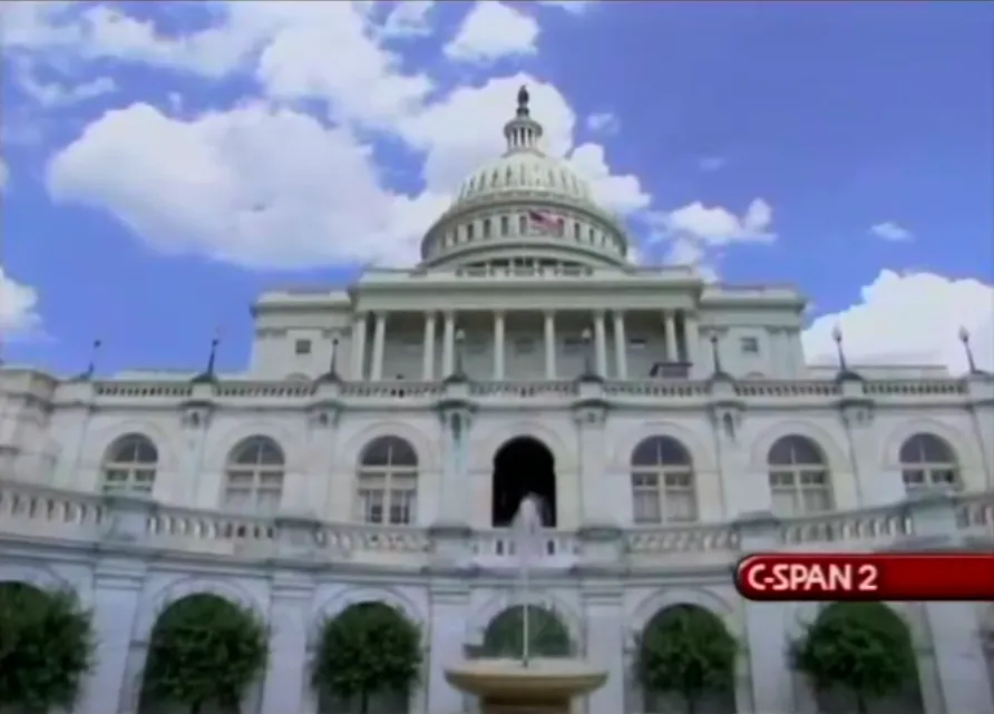 The United States Capitol building on a bright day with a blue sky and scattered clouds, viewed from a low angle; the C-SPAN 2 logo is visible in the lower right corner.