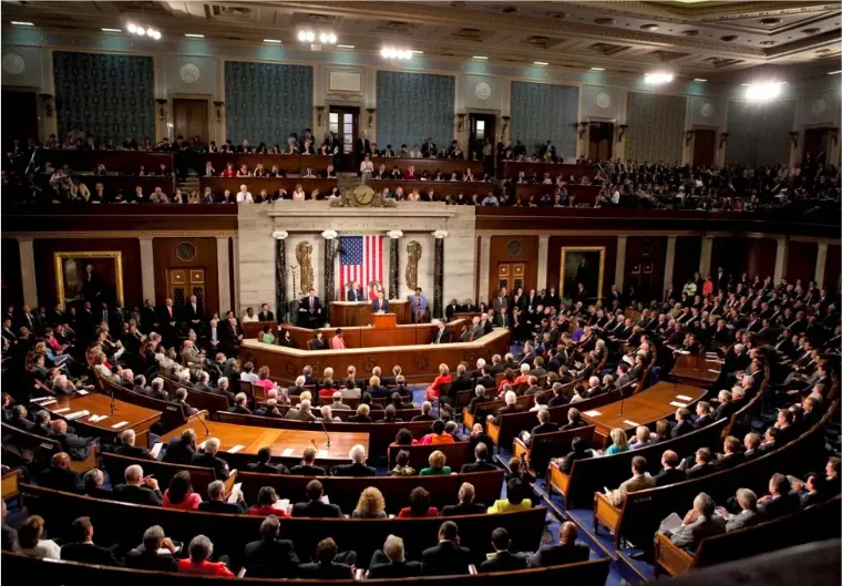 A wide-angle view of the U.S. House of Representatives chamber during a session, with members seated and an American flag displayed behind the speaker's podium.